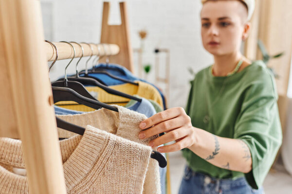 conscious decluttering, young tattooed woman sorting casual clothing on rack with hangers in modern living room at home, blurred background, sustainable fashion and mindful consumerism concept