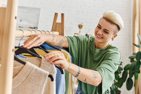 joyful tattooed woman with trendy hairstyle touching hangers on rack while sorting stylish casual clothes in living room at home, sustainable fashion and mindful consumerism concept