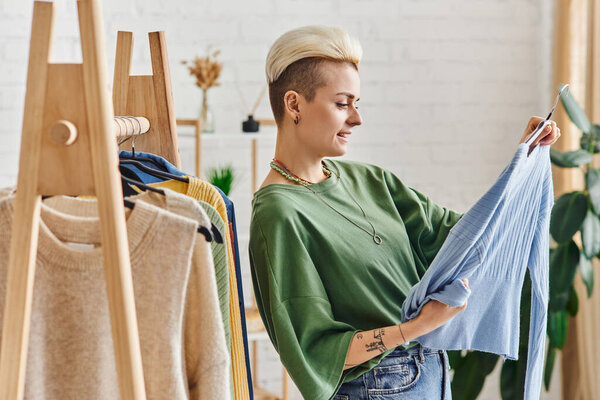 clothing sorting, side view of trendy and tattooed woman looking at blue cardigan near rack with wardrobe items on hangers, sustainable fashion and mindful consumerism concept