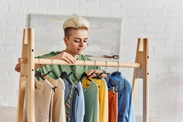 young and happy woman looking at fashionable casual clothes on rack in living room, thrift store finds, second-hand, conscious lifestyle, sustainable fashion and mindful consumerism concept