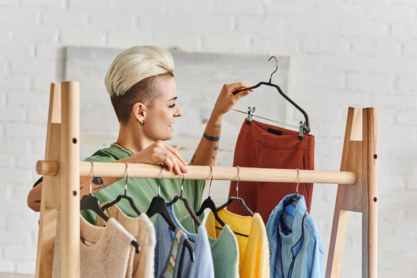 young and tattooed woman with trendy hairstyle holding hanger with trendy pants near rack with casual clothes in modern living room at home, sustainable fashion and mindful consumerism concept