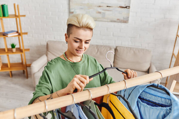 tattooed woman with happy smile and trendy hairstyle holding hanger near rack with clothing during decluttering process in living room, sustainable fashion and mindful consumerism concept
