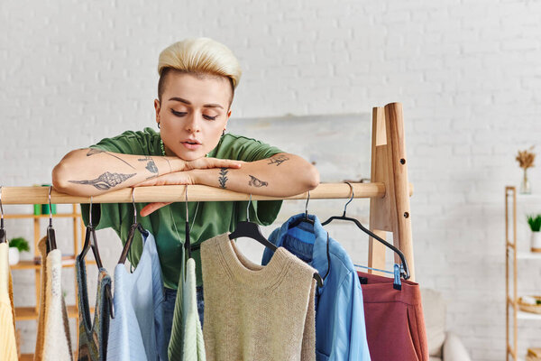 wardrobe items sorting, dreamy tattooed woman with trendy hairstyle leaning on rack with fashionable casual clothes on hangers, sustainable fashion and mindful consumerism concept