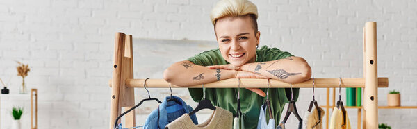 young tattooed woman with trendy hairstyle and happy face looking at camera and leaning on rack with hangers and stylish clothes at home, sustainable fashion and mindful consumerism concept, banner