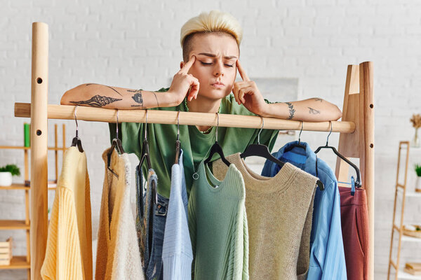 wardrobe items sorting, conscious decluttering, pensive tattooed woman touching head while thinking near rack with casual clothes at home, sustainable fashion and mindful consumerism concept