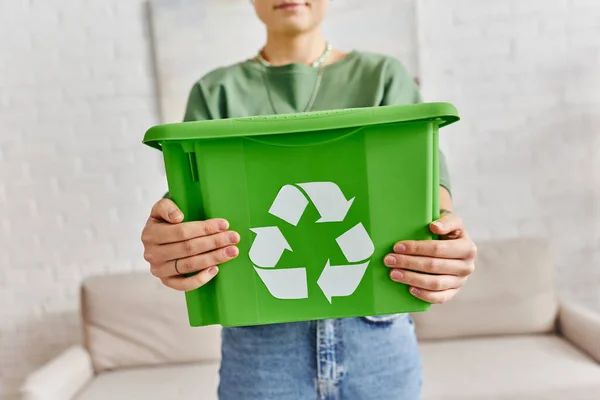 focus on green plastic box with recycling sign in hands of cropped woman standing at home on blurred background, sustainable living and environmentally friendly habits concept
