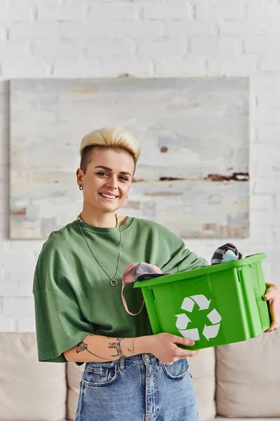 overjoyed tattooed woman with trendy hairstyle standing with garments in green recycling box and looking at camera at home, sustainable living and environmentally friendly habits concept