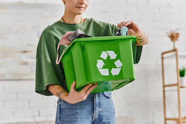 частичный взгляд на татуированную женщину, держащую зеленую коробку переработки с одеждой, стоя в гостиной, этическое потребление, устойчивая жизнь и экологически чистые привычки концепции