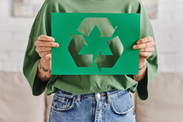 green living, partial view of young and tattooed woman in casual clothes holding green paper card with cutout recycled sign at home, sustainable and environmentally conscious lifestyle concept