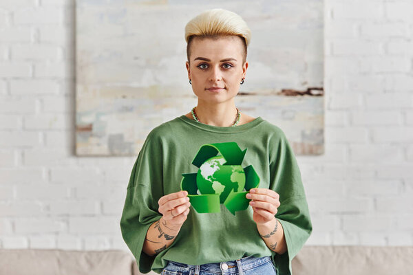 positive tattooed woman with trendy hairstyle showing green recycling sign with globe while smiling at camera in modern living room, sustainable living and environmental awareness concept