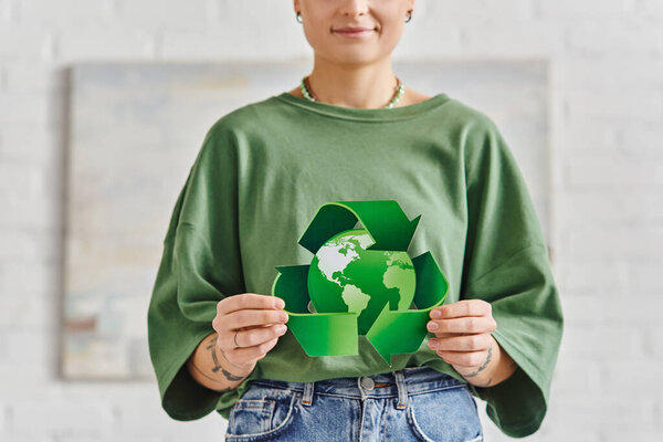 eco-conscious lifestyle, partial view of smiling tattooed woman in casual clothes holding green recycling symbol around globe at home, sustainable living and environmental awareness concept