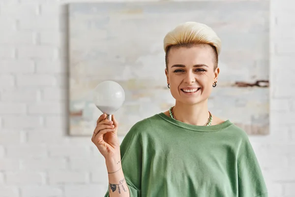 stock image ecological efficiency, joyful tattooed woman with trendy hairstyle holding energy saving light bulb and looking at camera in living room, sustainable lifestyle and environmentally conscious concept
