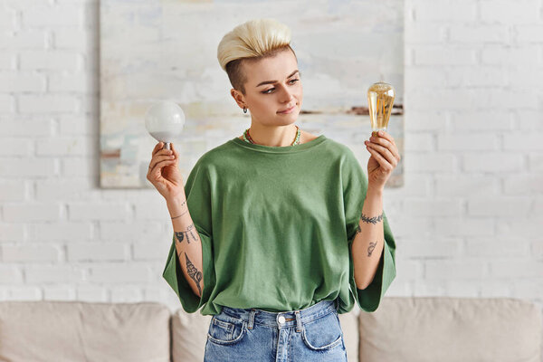 stylish tattooed woman in casual clothes, with trendy hairstyle comparing different energy saving light bulbs in modern living room, sustainable lifestyle and environmentally conscious concept
