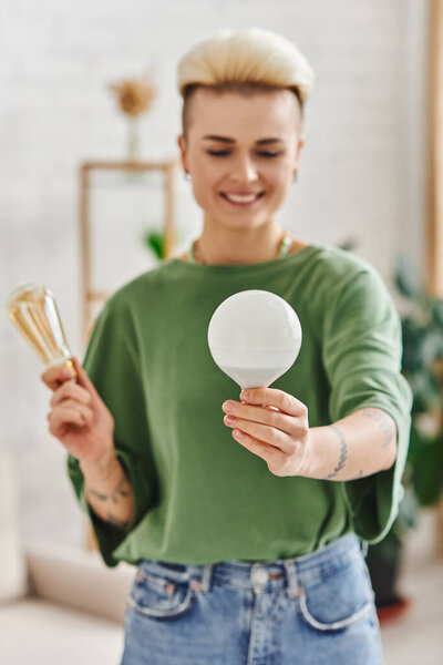 молодая и татуированная женщина в повседневной одежде, держа энергосберегающие лампочки и улыбаясь в гостиной на размытом фоне, устойчивом образе жизни и экологически сознательной концепции