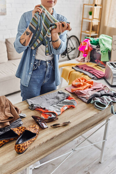 eco-friendly swaps, partial view of tattooed woman with knitted tank top near animal print shoes, sunglasses and casual clothes on table at home, sustainable living and mindful consumerism concept