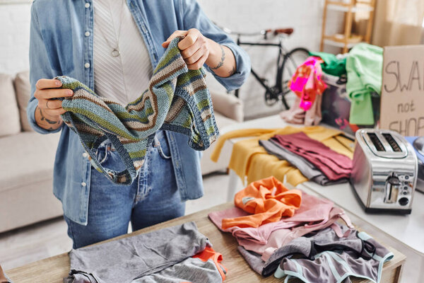 exchange market, partial view of tattooed woman sorting clothing and holding knitted tank top near table with pre-loved items and electric toaster, sustainable living and mindful consumerism concept