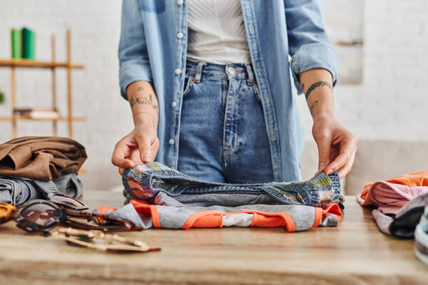 partial view of young and tattooed woman in casual clothes sorting wardrobe items on table in living room, exchange market, eco-friendly swaps, sustainable living and mindful consumerism concept