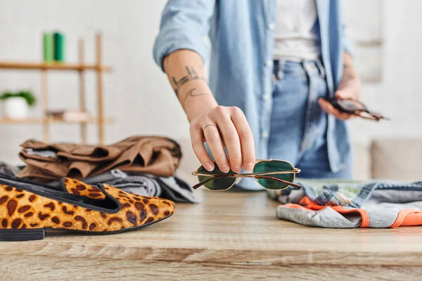 stock image cropped view of tattooed woman holding sunglasses near table with animal print shoes and second-hand clothes, resale market, sustainable living and mindful consumerism concept