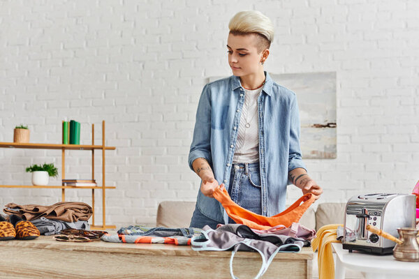 tattooed and casually styled woman sorting wardrobe items near electric toaster and cezve in modern living room, resale market, swap, sustainable living and promoting circular economy concept