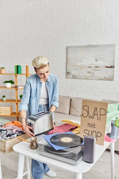 stock image casually styled and tattooed woman holding electric toaster near vinyl record player, cezve, pre-loved items and swap not shop card in living room, sustainable living and circular economy concept