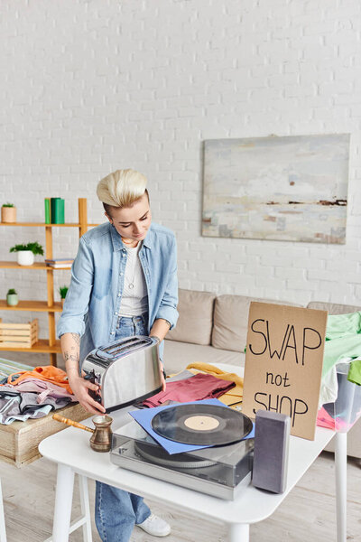 casually styled and tattooed woman holding electric toaster near vinyl record player, cezve, pre-loved items and swap not shop card in living room, sustainable living and circular economy concept