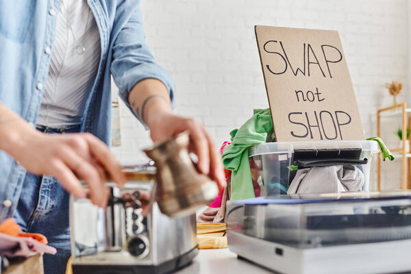 partial view of woman holding blurred cezve next to electric toaster, vinyl record player, plastic container with clothes and swap not shop card, sustainable living and circular economy concept