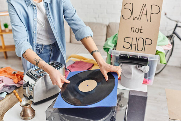 cropped view of tattooed woman holding vinyl disc near record player, electric toaster, cezve, plastic container with clothes and swap not shop card, sustainable living and circular economy concept