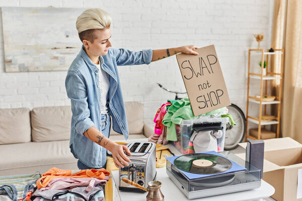 tattooed woman with trendy hairstyle holding swap not shop card near vinyl record player, electric toaster, cezve and wardrobe clothes in living room, sustainable living and circular economy concept