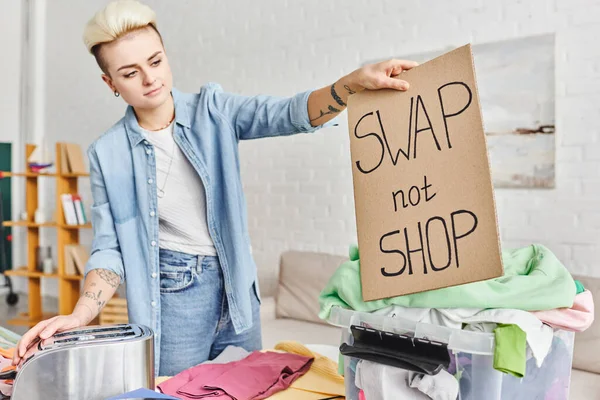 tattooed woman in casual clothes holding swap not shop card near electric toaster, plastic container and clothes in modern living room, sustainable living and circular economy concept
