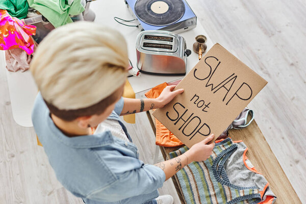 top view of young tattooed woman holding card with swap not shop lettering near clothes, electric toaster, vinyl record player and cezve, sustainable living and circular economy concept