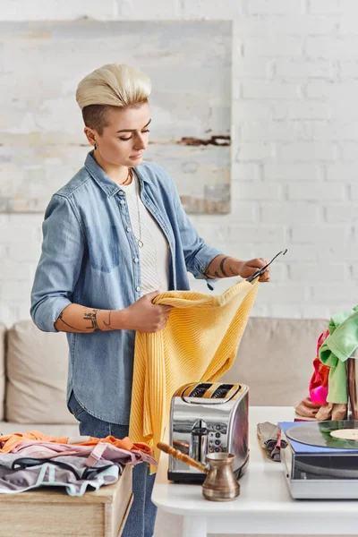 stock image exchange market, tattooed woman with trendy hairstyle holding yellow jumper near table with electric toaster, vinyl record player and clothes, sustainable living and mindful consumerism concept