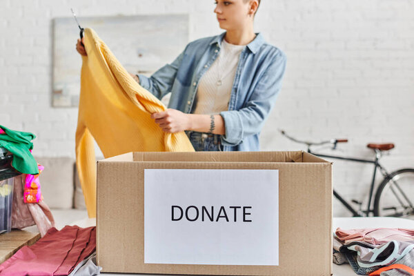 татуированная женщина в повседневной одежде смотрит на желтый свитер возле гардероба одежды и коробки с пожертвованием буквы, размытый фон, устойчивая жизнь и социальная ответственность