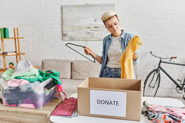 татуированная женщина с модной прической, держащая вешалку и желтый свитер рядом с пластиковым контейнером с одеждой и коробкой для пожертвований в гостиной, концепцией устойчивой жизни и социальной ответственности