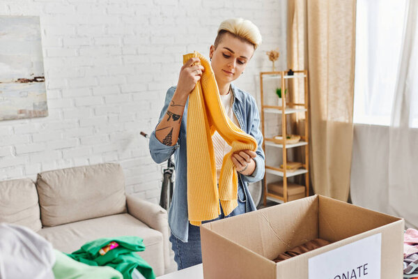 volunteering and charity, trendy and tattooed woman standing with yellow jumper near carton box while sorting clothing for donation at home, sustainable living and social responsibility concept