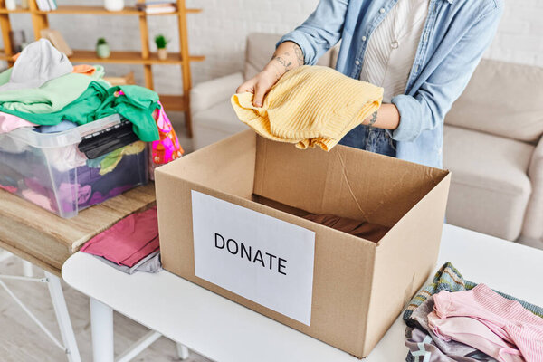 частичный взгляд на молодую и татуированную женщину, держащую желтый свитер над коробкой с надписью донора возле пластикового контейнера с одеждой, концепцией устойчивой жизни и социальной ответственности