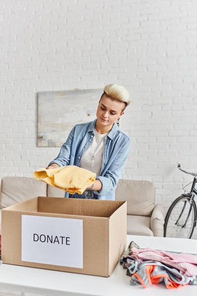charity and volunteering, young tattooed woman holding yellow jumper near wardrobe items and carton box with donate lettering, sustainable living and social responsibility concept