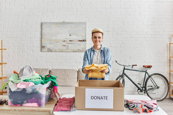 молодая и позитивная женщина с модной прической улыбается перед камерой и держит желтый свитер рядом с коробкой пожертвований и пластиковым контейнером с одеждой, устойчивой жизни и социальной ответственности