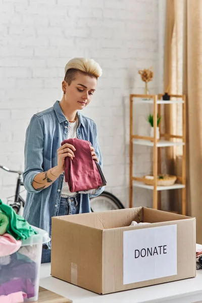 donating for a cause, tattooed woman with trendy hairstyle holding clothes near carton box with donate lettering in modern living room, sustainable living and social responsibility concept