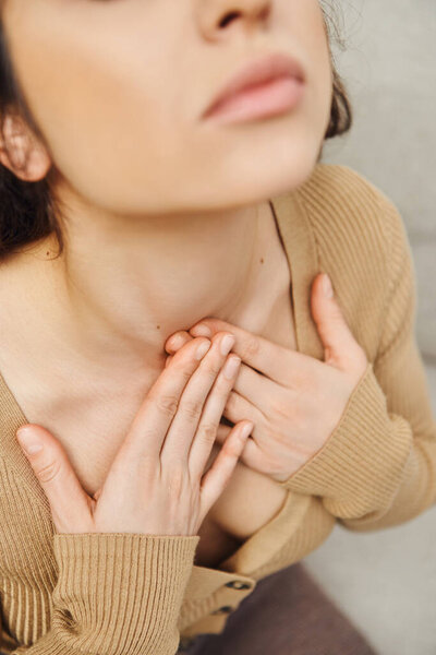 High angle view of young brunette woman in casual jumper touching neck during self-massage of thyroid gland in blurred house, self-care ritual and holistic wellness practices concept, tension relief