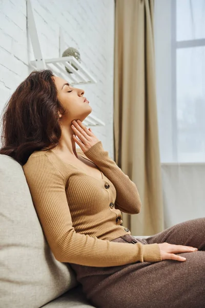 stock image Side view of relaxed and young brunette woman in brown jumper touching neck during self-massage for lymphatic system on couch at home, self-care ritual and holistic wellness practices concept