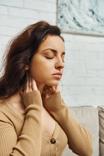 Portrait of young woman in casual brown jumper massaging lymphatic nodes and relaxing while sitting on blurred couch at home, self-care ritual and holistic wellness practices concept, tension relief