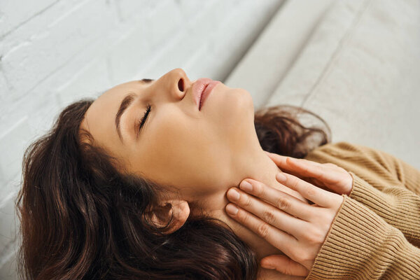 Close up view of relaxed brunette woman in brown jumper massaging neck during lymphatic drainage support and sitting on couch at home, self-care ritual and holistic healing concept, tension relief