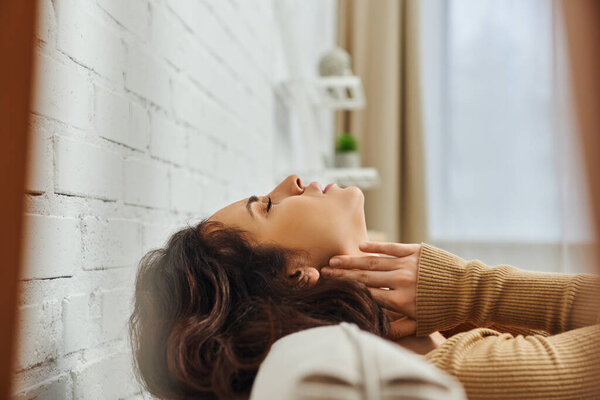 Side view of relaxed brunette woman in brown jumper massaging lymphatic nodes on neck during nurturing self-care on couch at home, self-care ritual and holistic healing concept, tension relief