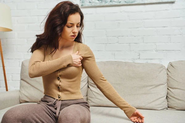 Young brunette woman in brown jumper suffering from pain while massaging lymphatic nodes on armpit and sitting on couch at home, self-care ritual and holistic wellness practices concept