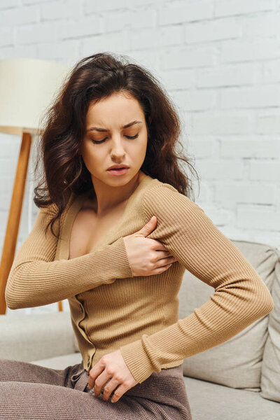 Портрет юной брюнетки в непринужденном коричневом свитере, массирующей лимфатические узлы подмышки во время внимательного ухода за собой на диване дома, повышения самосознания и расслабления тела