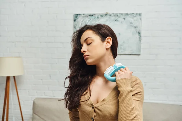 Young brunette woman in brown jumper holding handled massager and massaging lymphatic nodes on neck in blurred house, enhancing self-awareness and body relaxation concept, balancing energy
