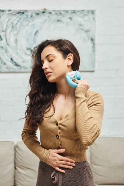 stock image Young brunette and relaxed woman in brown jumper massaging lymphatic nodes with handled massager while standing in blurred house, self-care ritual and holistic wellness practices concept