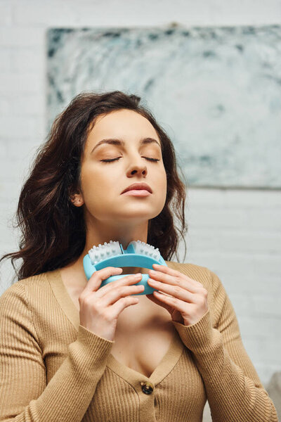Portrait of young woman with closed eyes in casual brown jumper massaging neck and lymphatic nodes with handled massager at home, enhancing self-awareness and body relaxation concept 