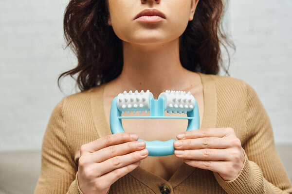 Обрезанный вид юной брюнетки в непринужденном коричневом джампере с массажером для самомассажа и стоя дома, повышение самосознания и расслабления тела, балансирование энергии