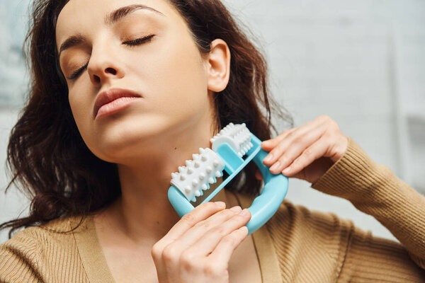 Портрет брюнетки с закрытыми глазами в коричневом свитере и массирующей лимфатической системе на шее с ручным массажером дома, улучшающим самосознание и концепцию расслабления тела